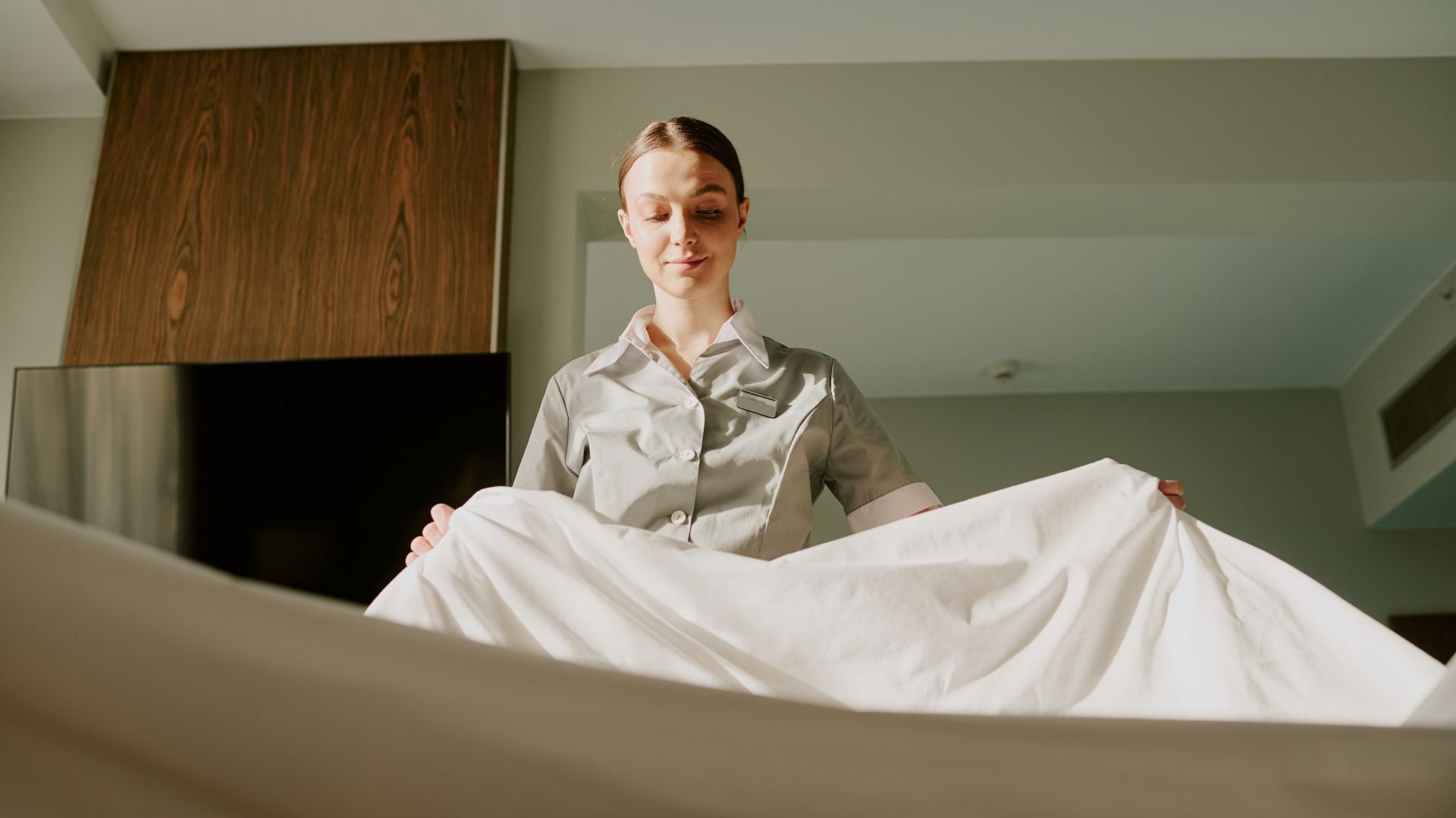 Hotel housekeeper making bed with white linens in sunlit room