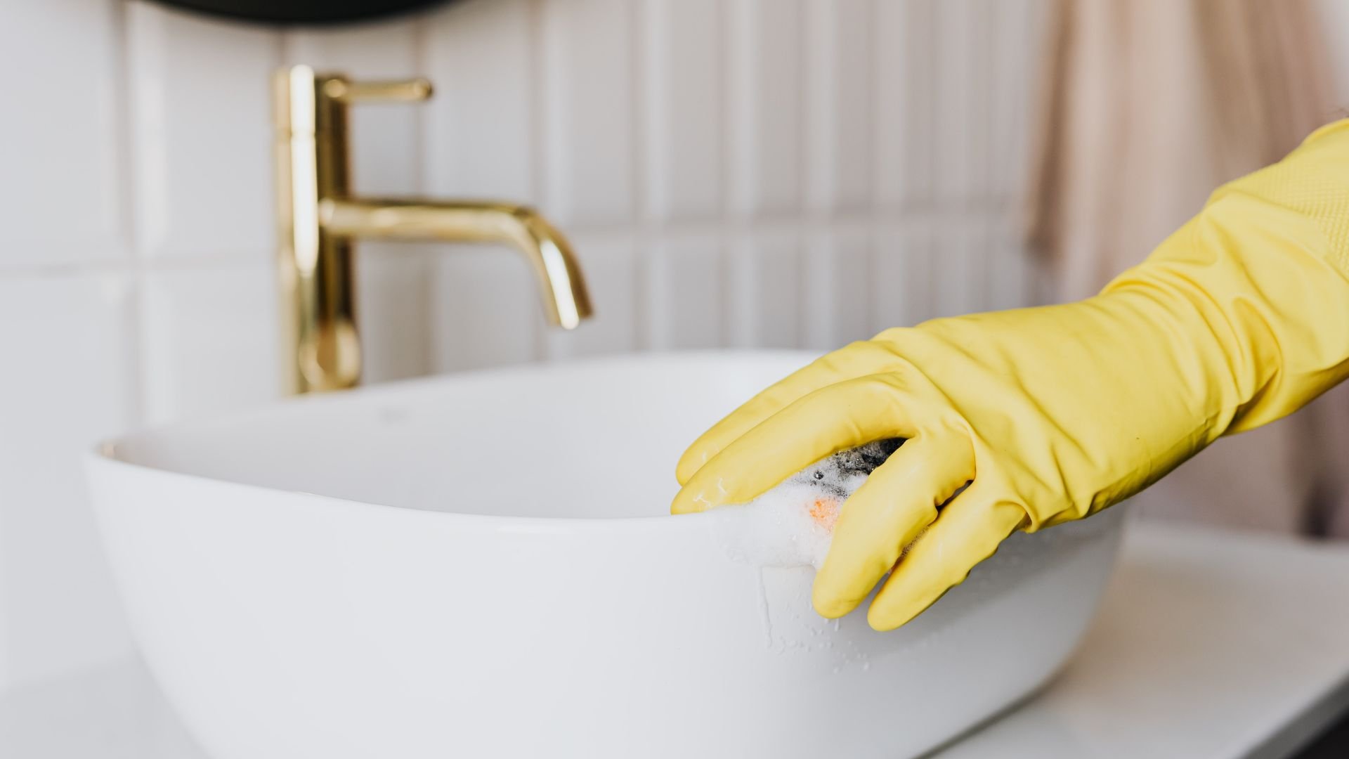 Yellow gloved hand cleaning white sink with golden faucet